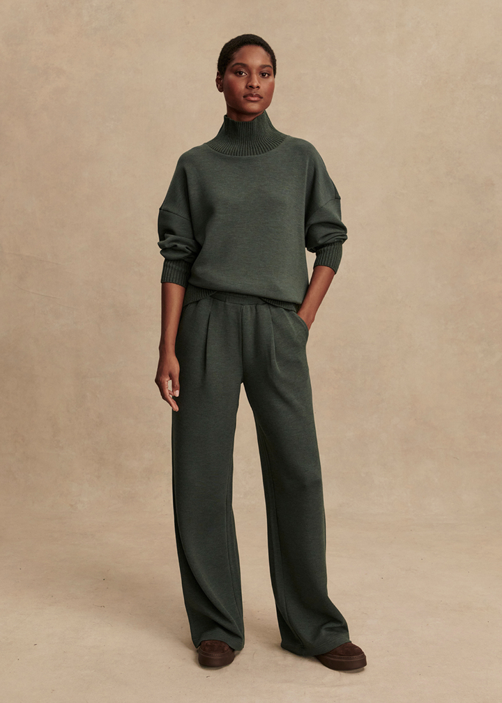 Woman wearing a dark green turtleneck and wide leg pants in front of a tan background with hand in pocket