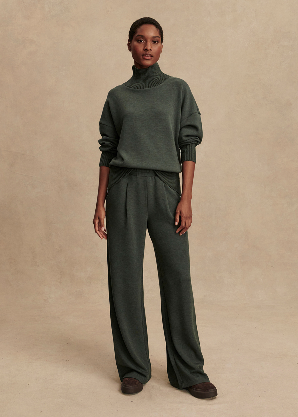 Woman wearing a dark green turtleneck and wide leg pants in front of a tan background