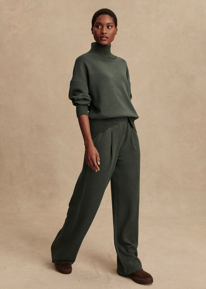Woman walking wearing a dark green turtleneck and wide leg pants in front of a tan background