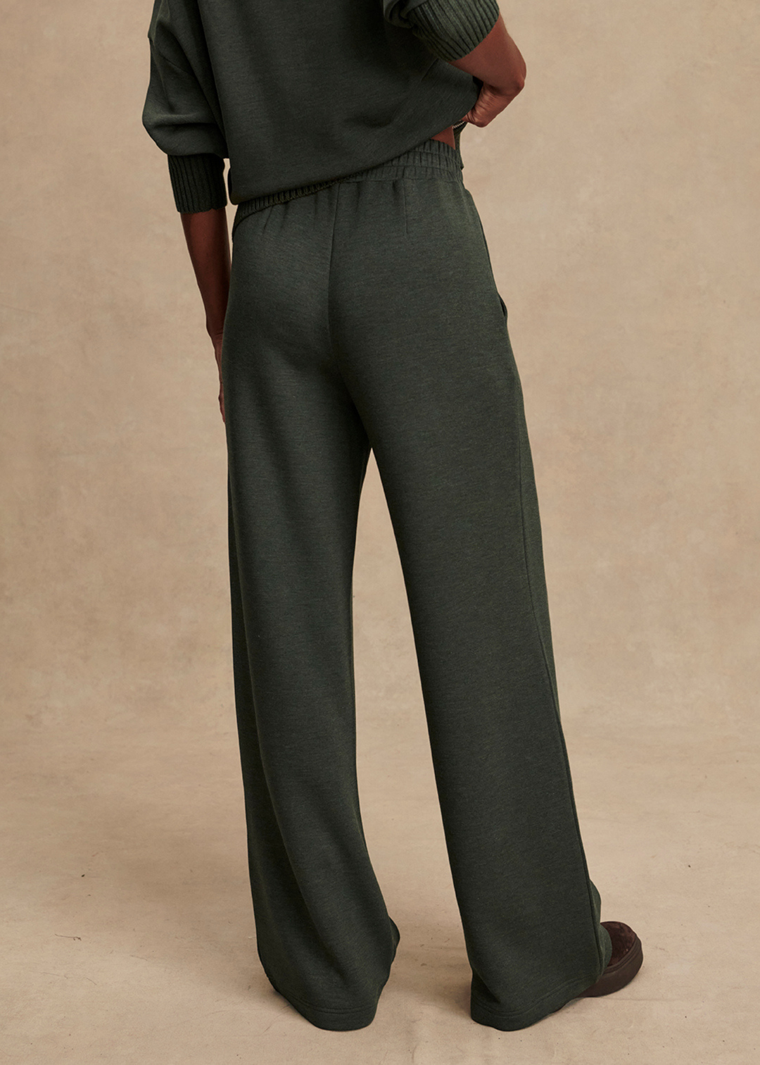 Woman turned around wearing dark green wide leg pants in front of a tan background