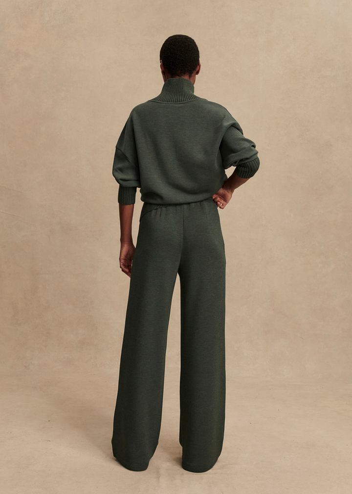 Woman turned around wearing dark green wide leg pants and turtleneck in front of tan background