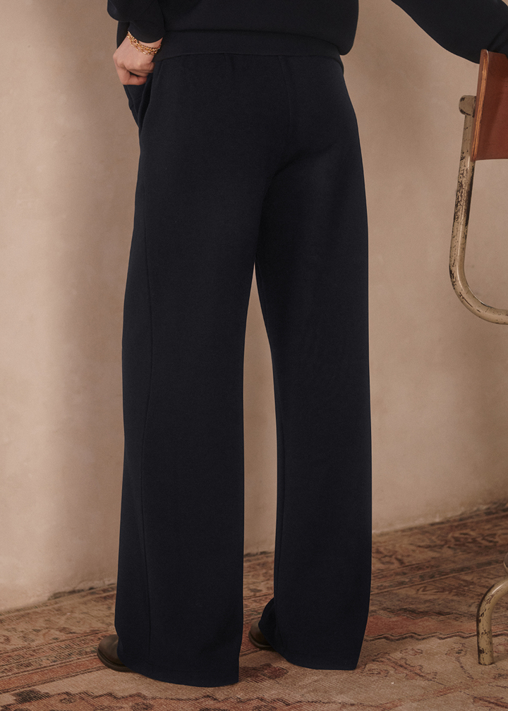 Close up of a woman turned around wearing wide leg navy sweatpants with a matching top in front of a tan background