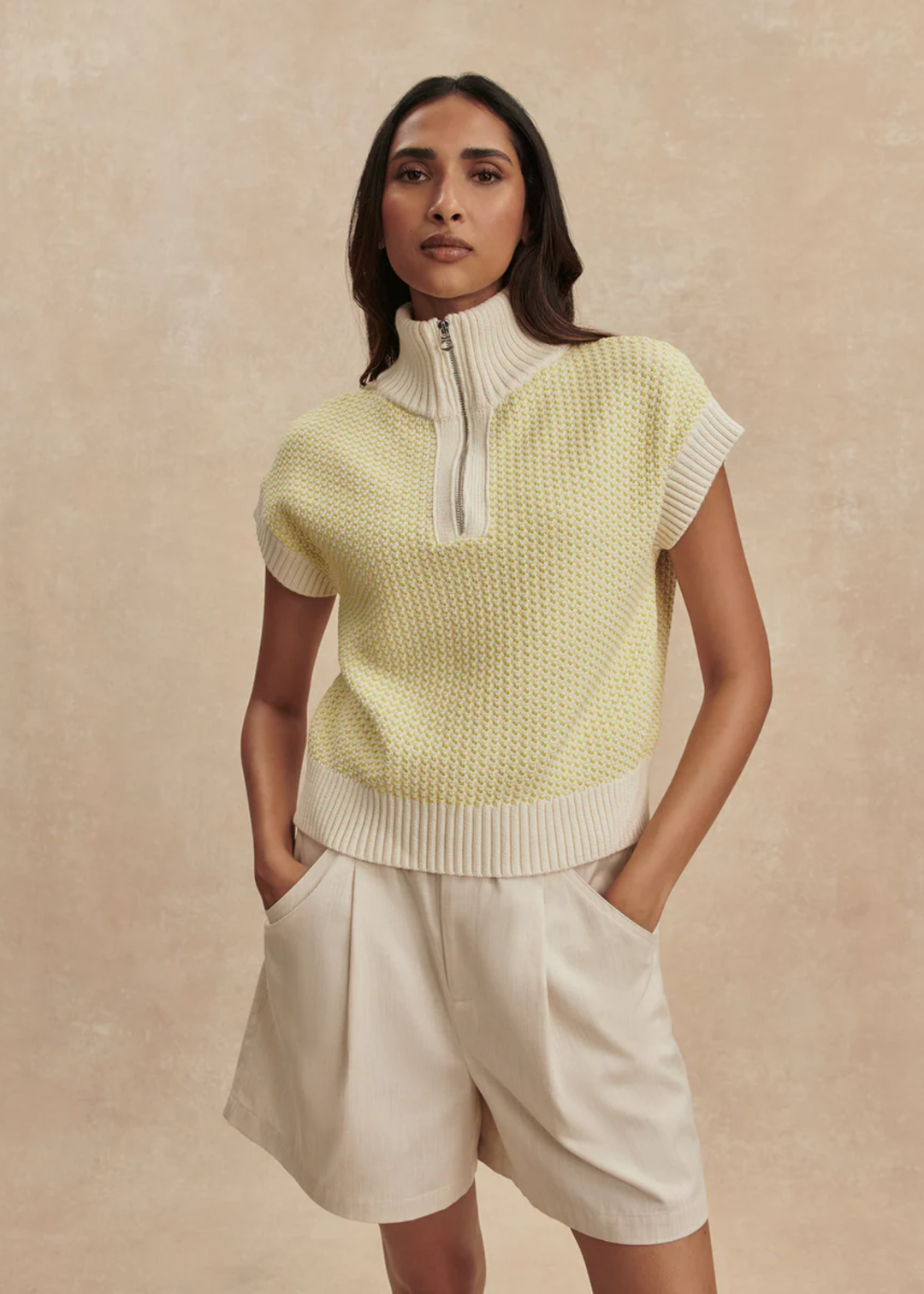 Woman wearing a yellow knit top with cream shorts against a tan background
