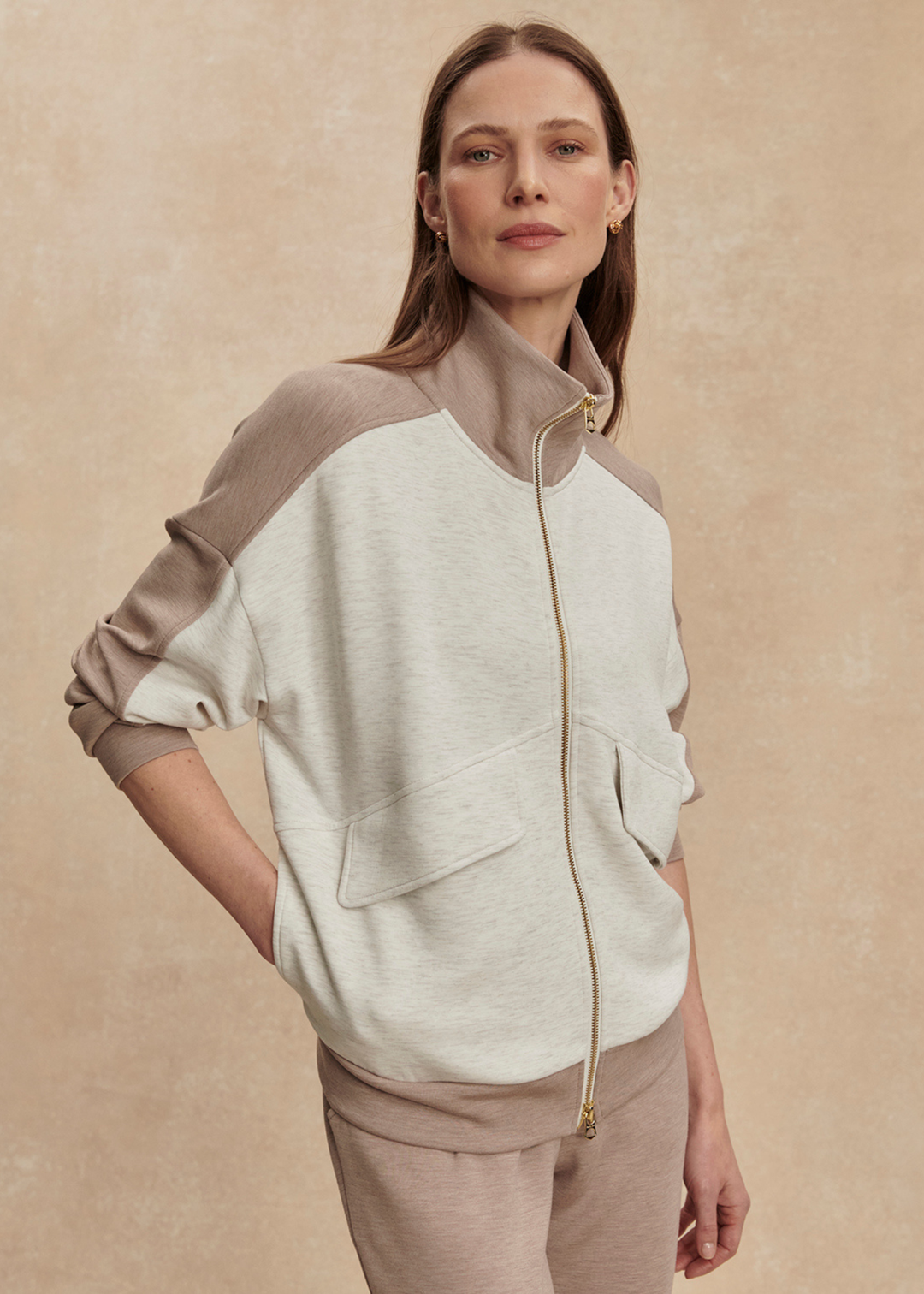 Woman wearing a taupe and white zip up sweatshirt with taupe pants against a tan background