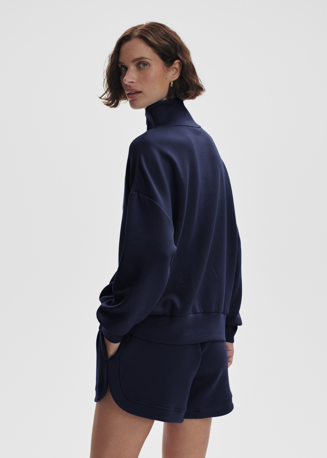 Woman turned to the side wearing a navy half zip sweatshirt top with matching shorts in front of a white background