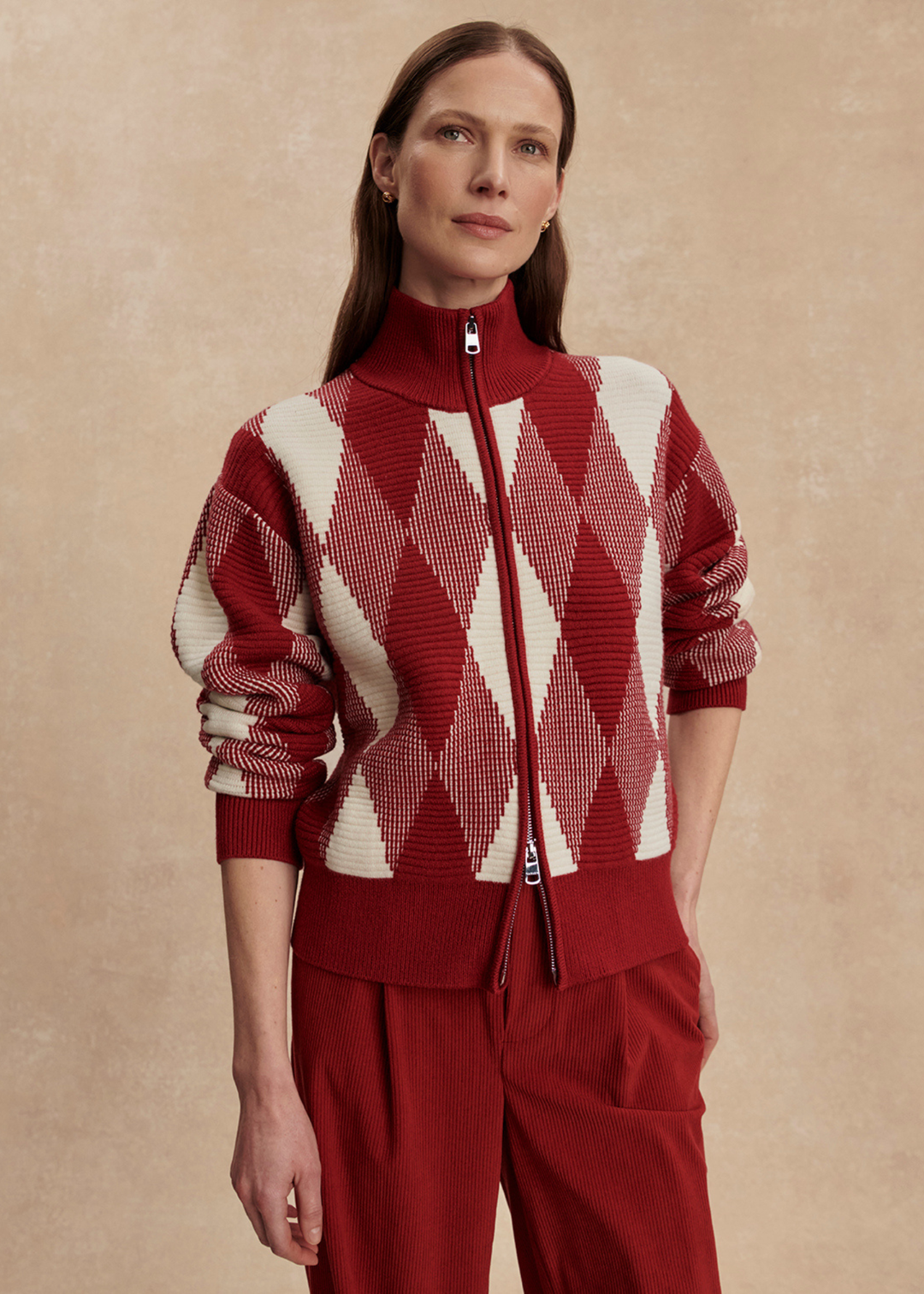 Woman wearing a red and white patterned sweater with red pants with hand in pocket against a tan background