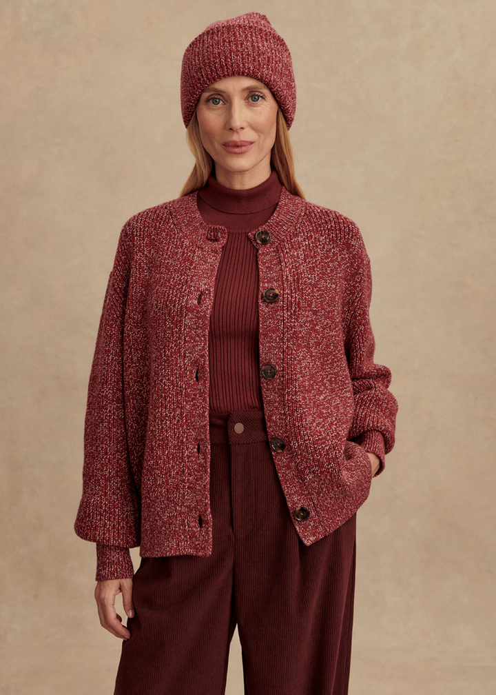 Woman wearing a long sleeve ribbed button up cardigan in a red color with a white fleck pattern with other red clothing in front of a tan background