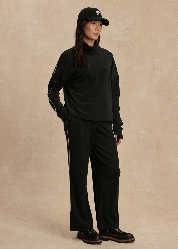 Woman with hand in pocket wearing a black set with white trim against a tan background