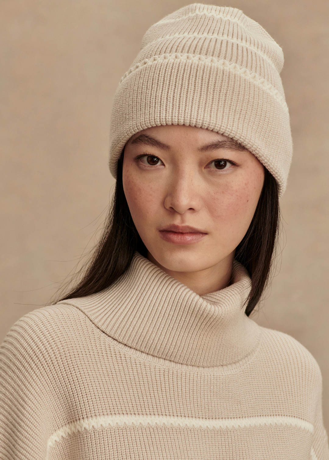 Woman wearing a tan and white striped beanie with a matching sweater in front of a tan background