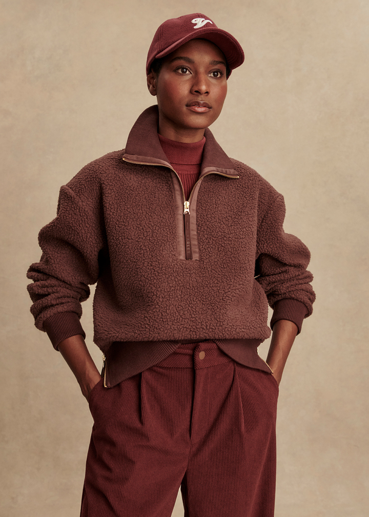Woman wearing a maroon half zip fleece pullover with maroon pants in front of a tan background