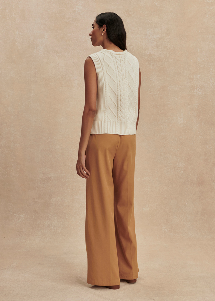 Woman turned around wearing a cream cable knit sweater vest with brown pants