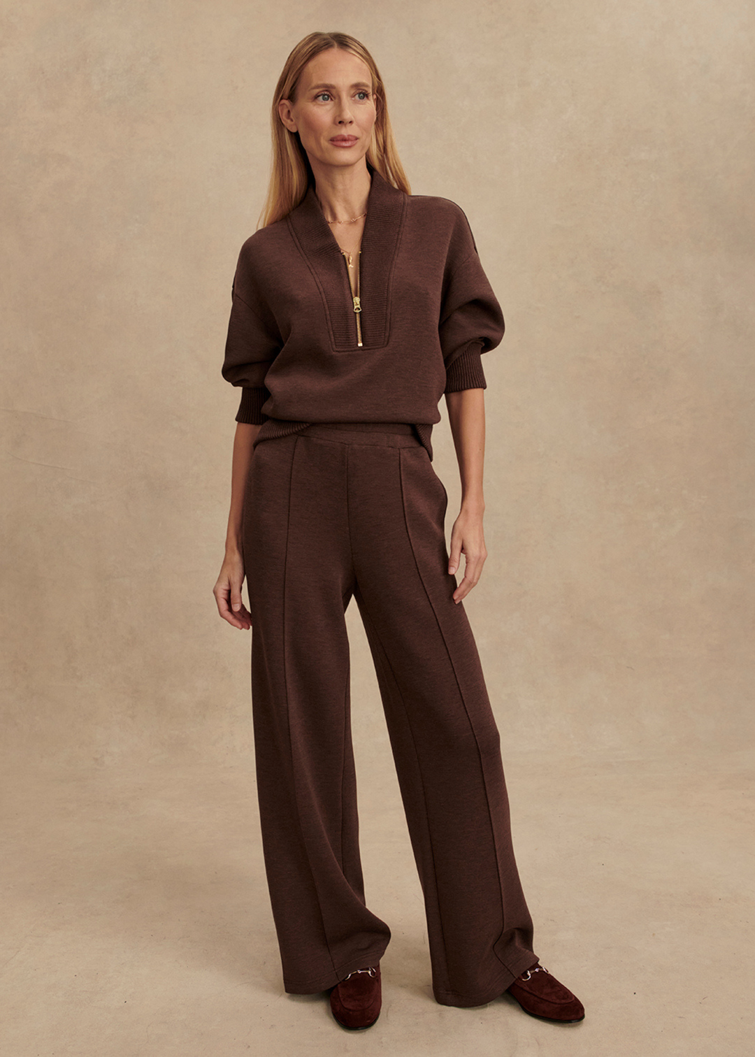Woman wearing brown wide leg pants with a matching half zip sweatshirt in front of a tan background