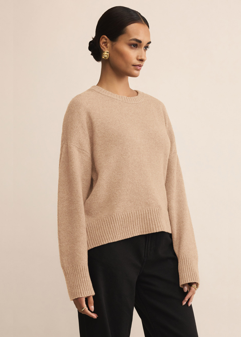Woman turned to the side wearing a tan sweater with dark jeans standing in front of a white background