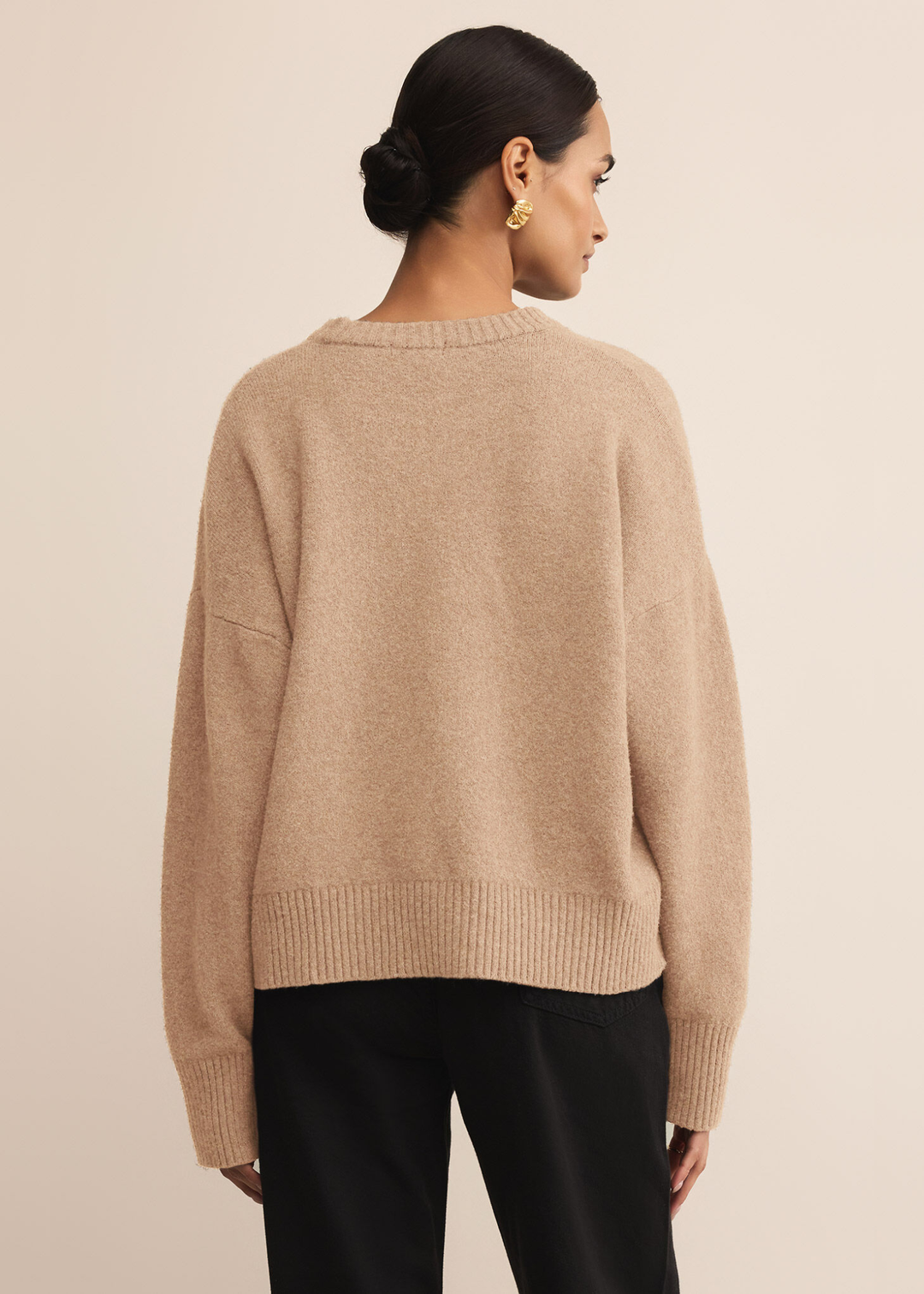 Woman turned around wearing a tan sweater with dark jeans standing in front of a white background