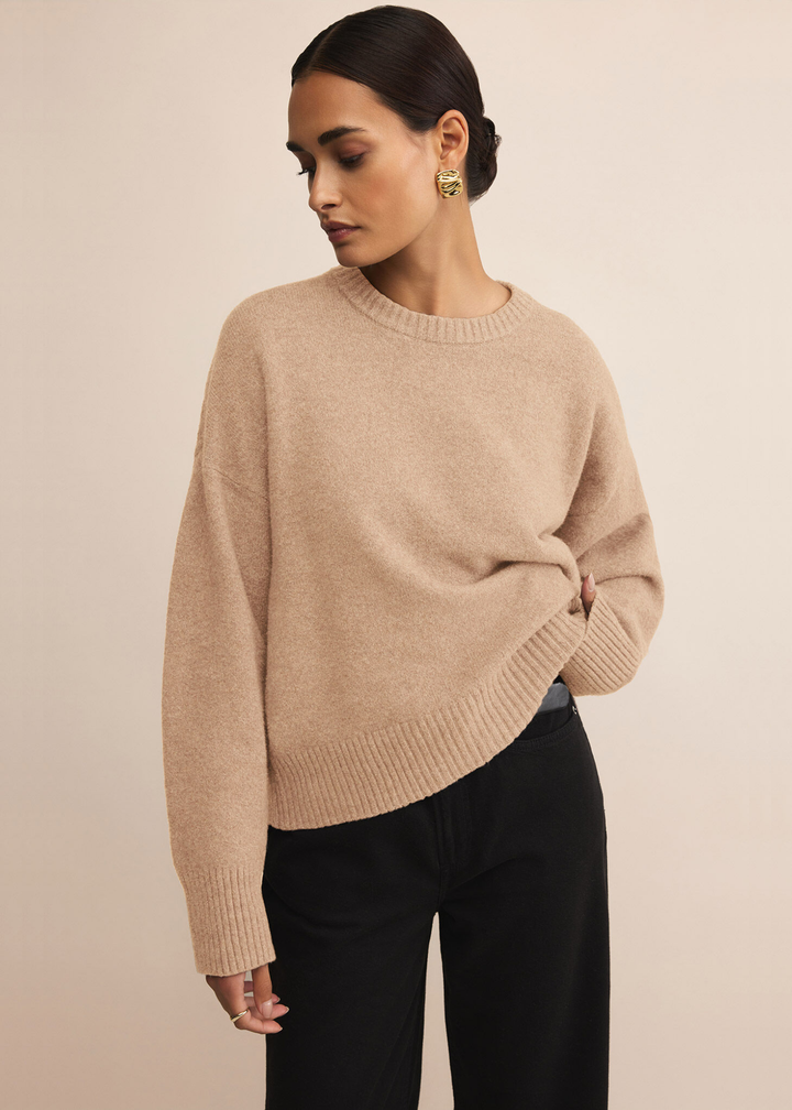 Woman wearing a tan sweater with dark jeans standing in front of a white background