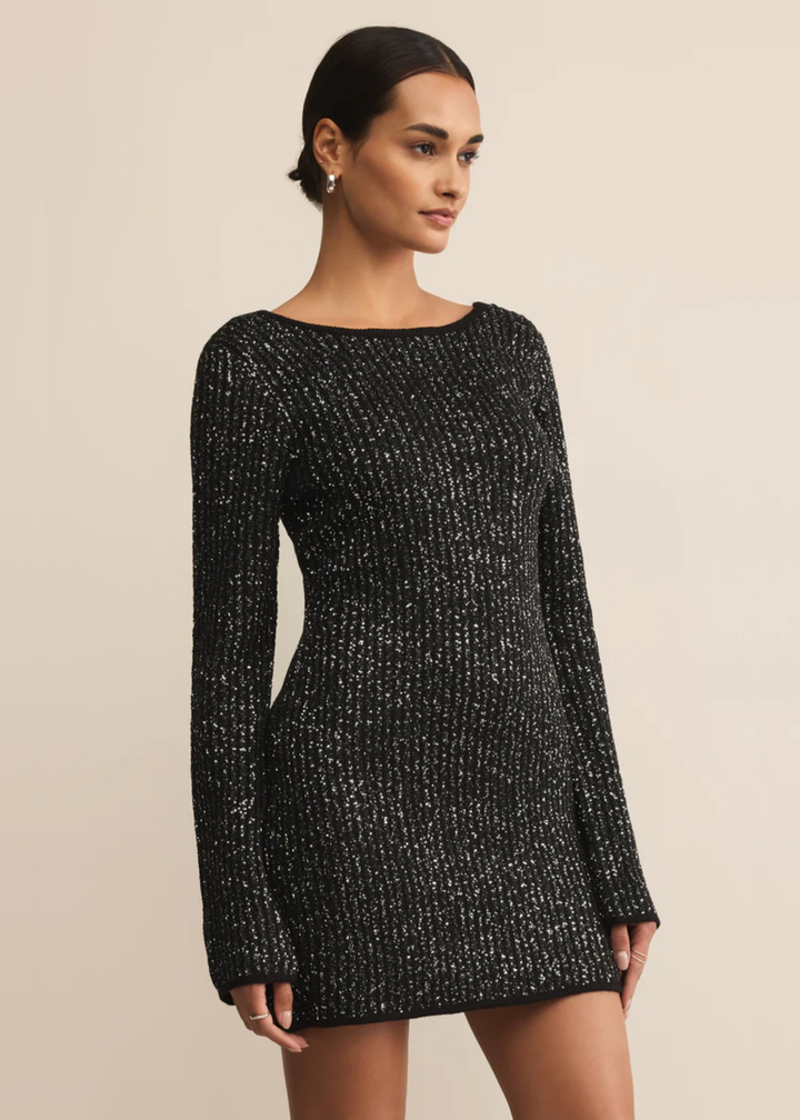 Woman turned to the side wearing a black sequin mini dress with tall black boots in front of a tan background