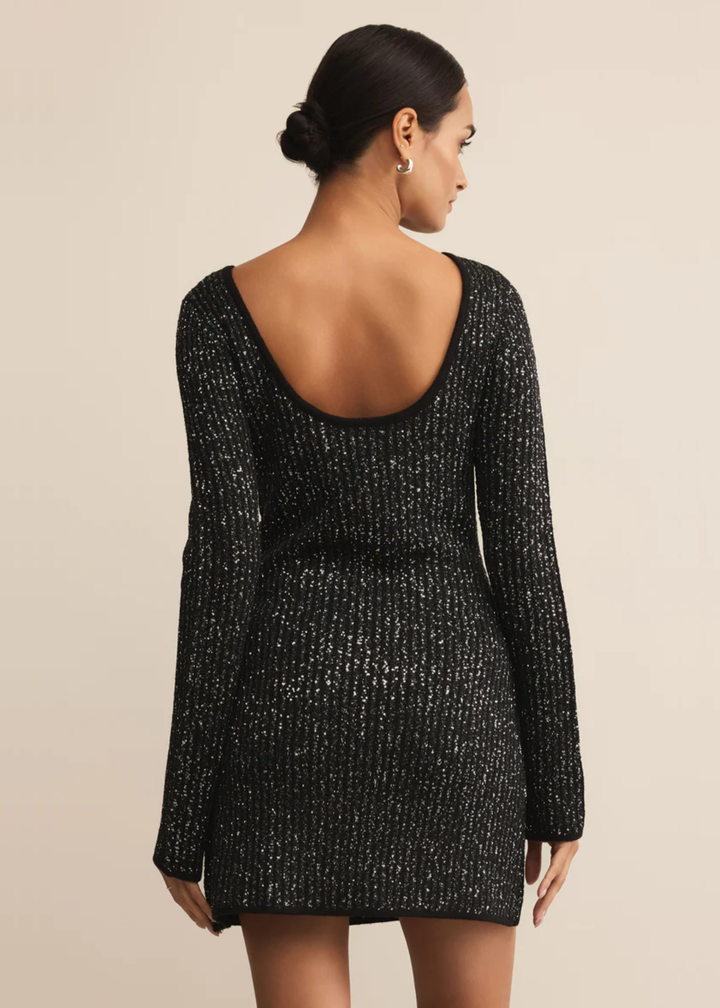 Woman turned around wearing a black sequin mini dress with tall black boots in front of a tan background