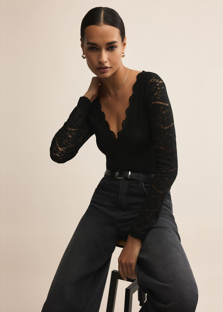 Woman sitting on a stool wearing a black lace bodysuit with scalloped trim and black jeans against a tan background