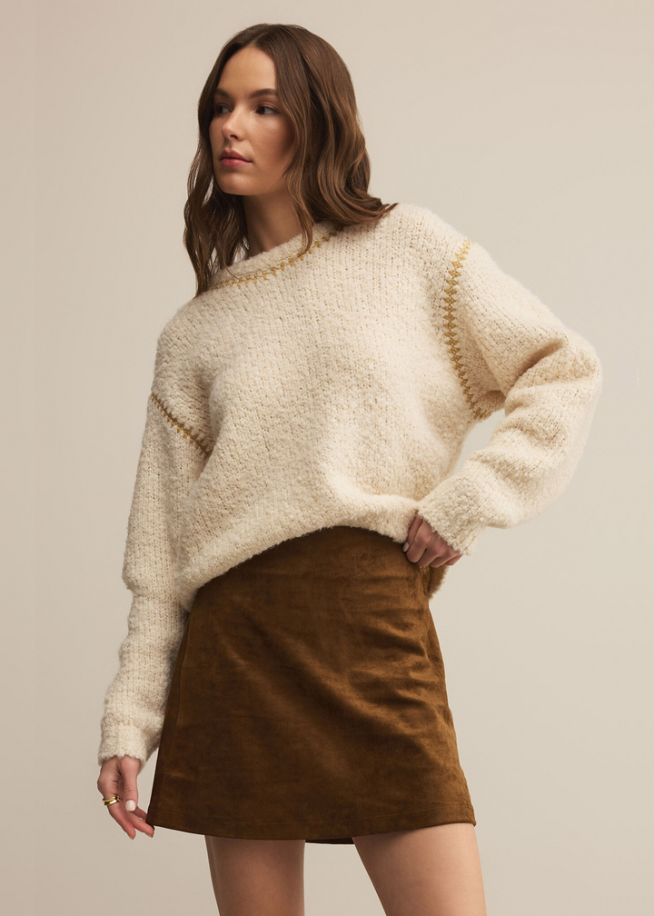 Woman wearing a cream colored sweater with brown stitching in front of a grey background