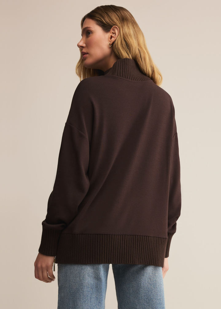 Woman turned around wearing a brown sweater with a mock neckline and jeans standing in front of a tan background