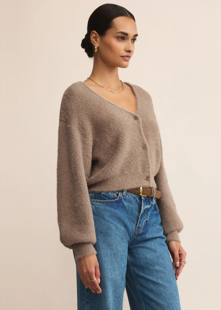 Woman turned to the side wearing a fuzzy light brown cardigan with jeans in front of a grey background