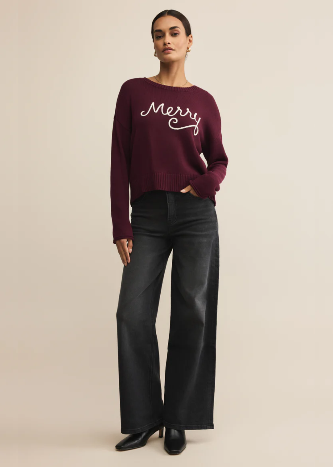 Woman wearing a maroon sweater with white "Merry" lettering and black jeans and boots in front of a tan background