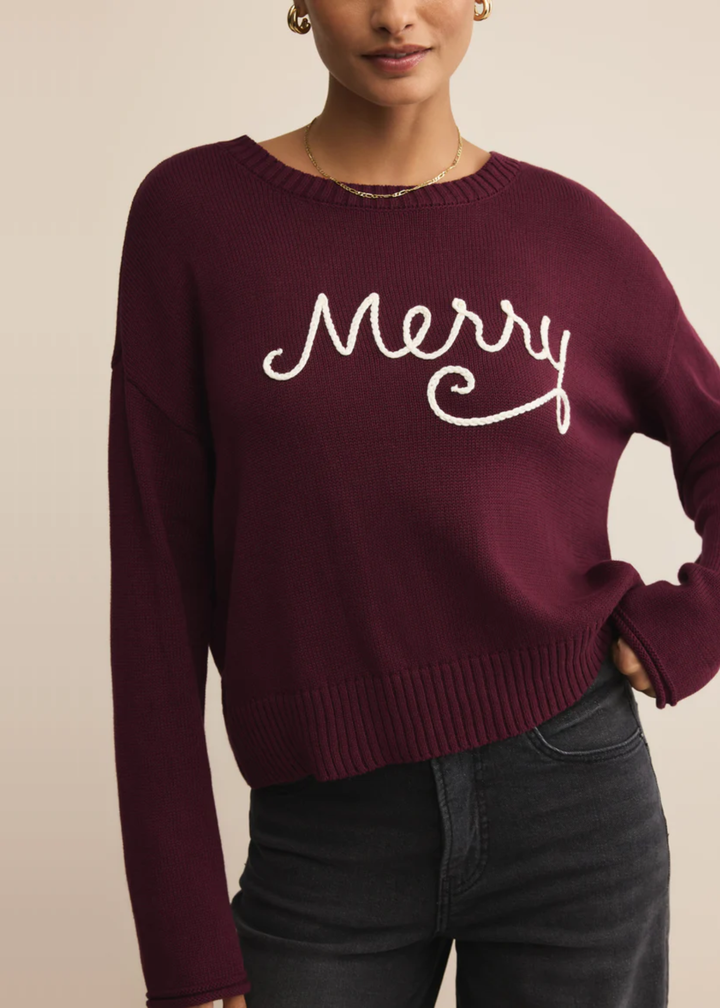 Woman with hand in pocket wearing a maroon sweater with white "Merry" lettering in front of a tan background