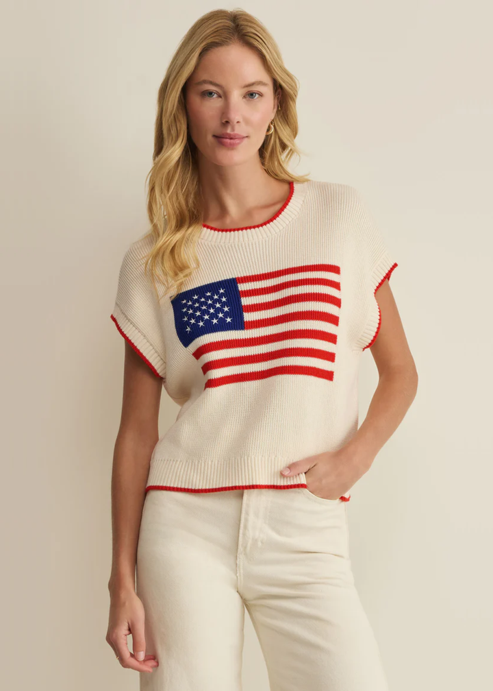Woman wearing a cream sleeveless sweater with an American flag graphic and white jeans