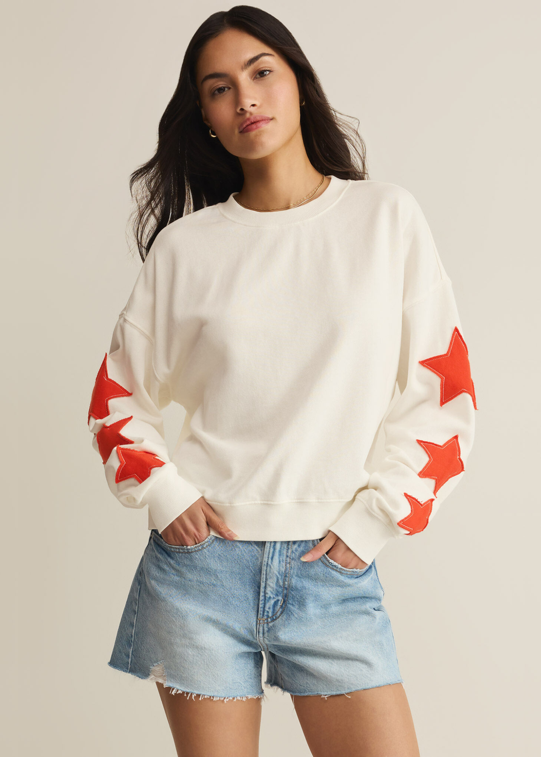 Woman wearing a cream sweatshirt with red stars stitched on the sleeves with denim shorts