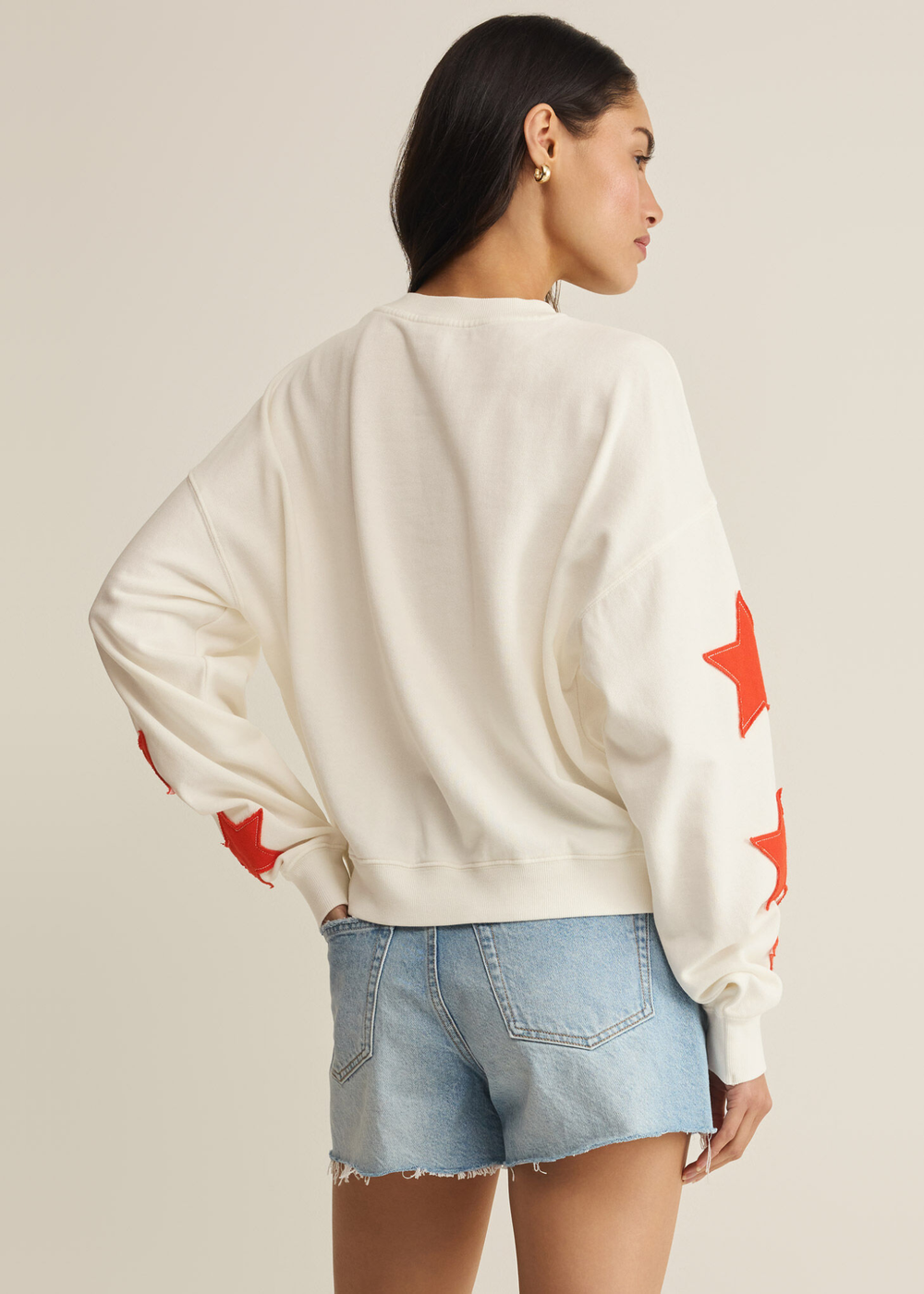 Woman turned around wearing a cream sweatshirt with red stars stitched on the sleeves with denim shorts