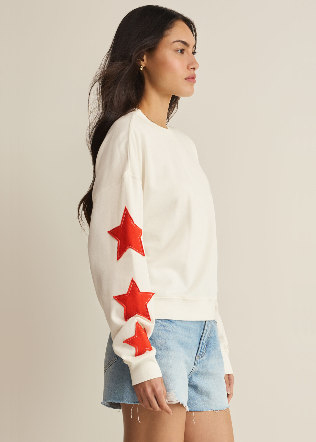 Woman turned to the side wearing a cream sweatshirt with red stars stitched on the sleeves with denim shorts