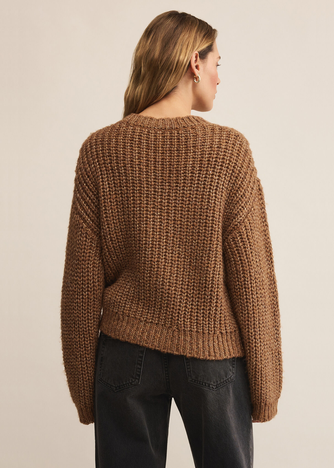 Woman turned around wearing a brown sweater with gold flecks with black jeans standing in front of a white background