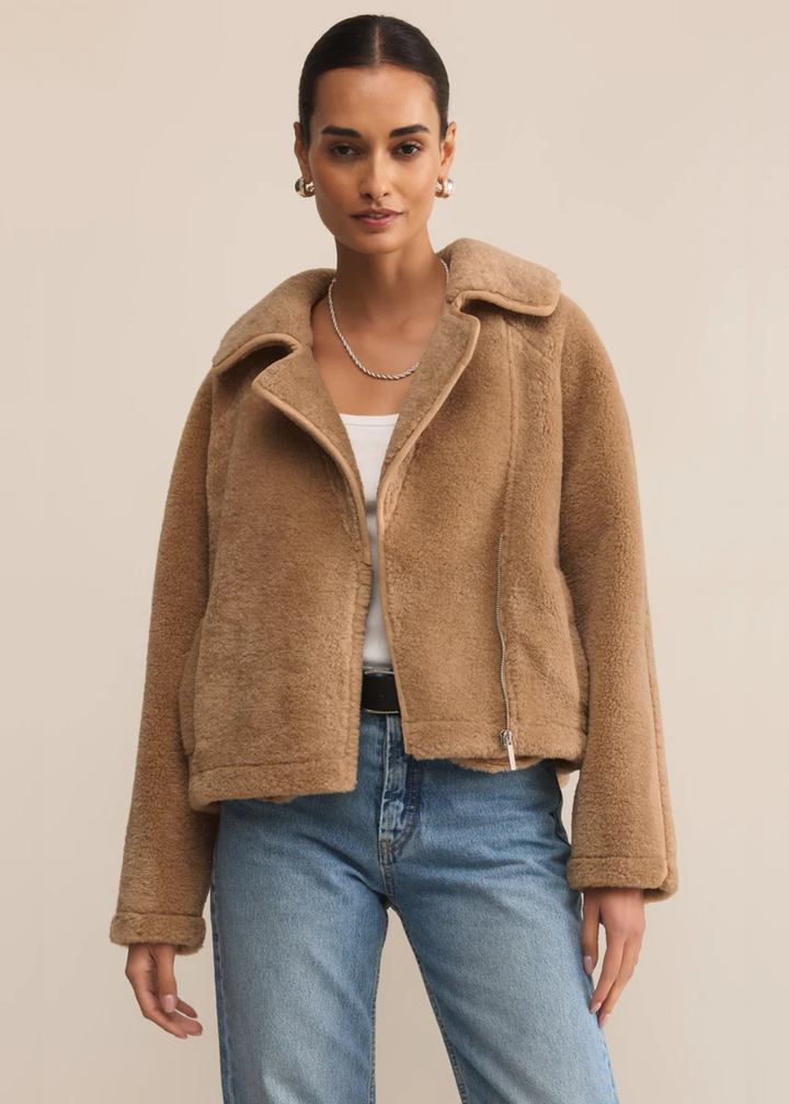 Woman wearing a brown sherpa jacket over a white shirt and jeans standing in front of a tan background