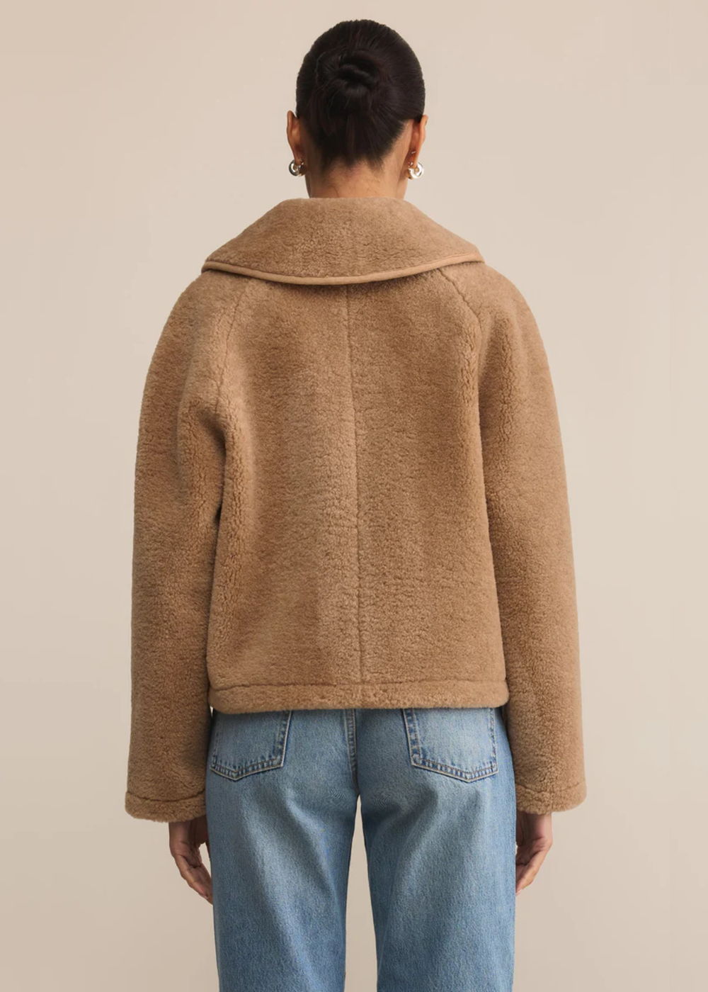 Woman turned around wearing a brown sherpa jacket over a white shirt and jeans standing in front of a tan background