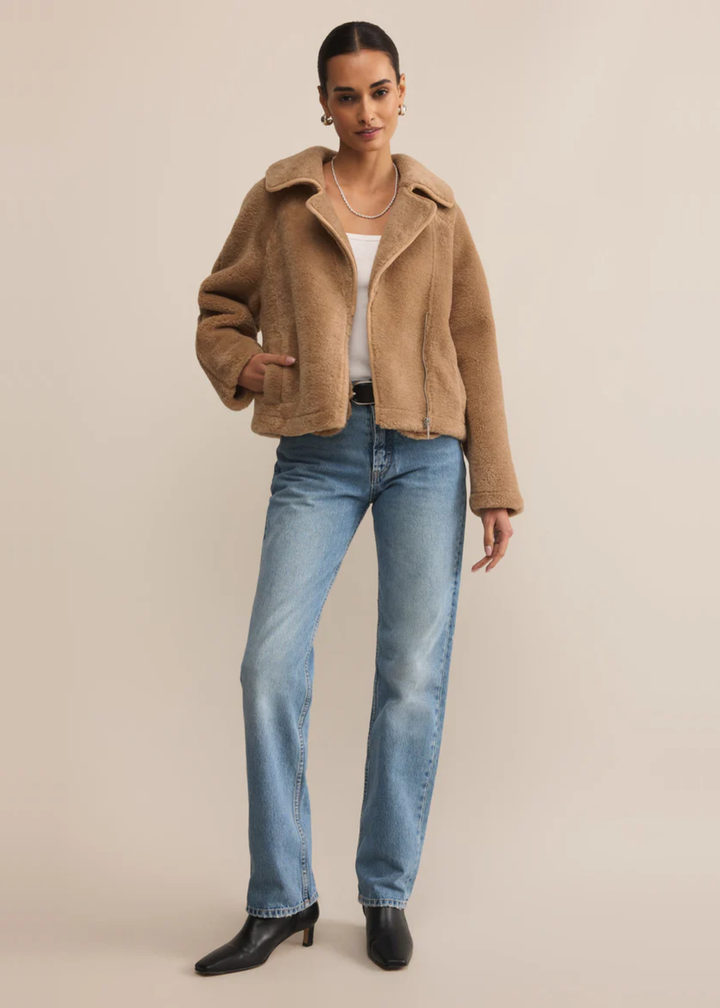 Woman wearing a brown sherpa jacket over a white shirt and jeans standing in front of a tan background