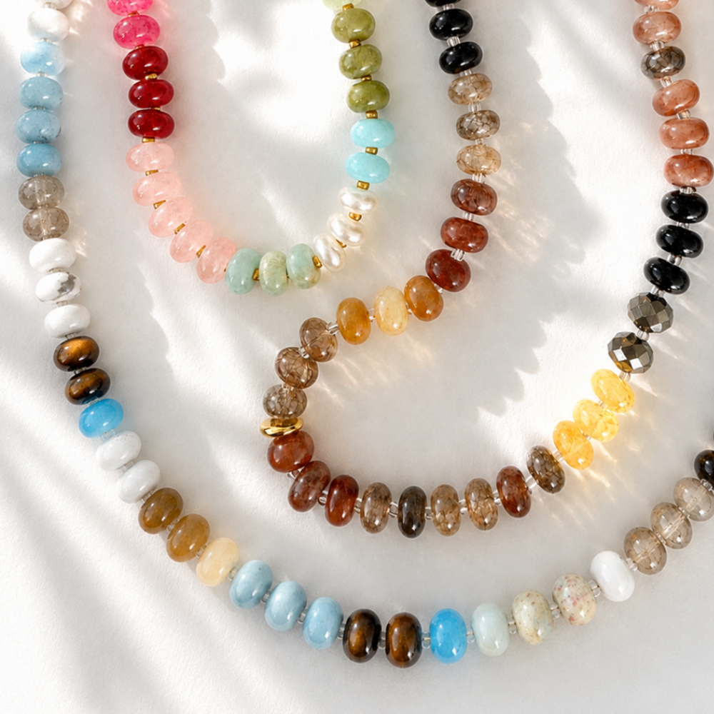Four beaded necklaces arranged in overlapping loops on a white fabric background. The necklaces feature smooth, polished round beads in various colors including light blue, white, brown, pink, mint green, orange, yellow, black, and smoky gray, with some strands showing small gold spacers.
