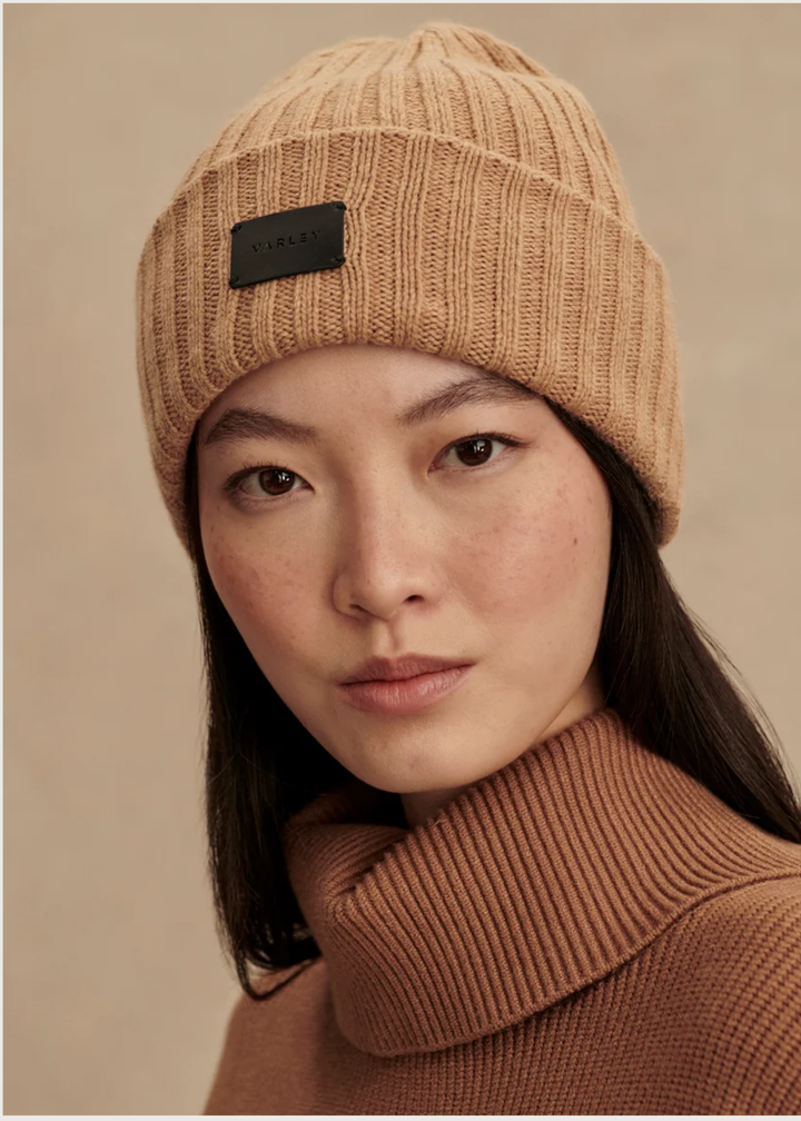 A close-up portrait of a woman wearing a ribbed knit beanie in a warm tan/camel color with a small black brand patch on the front, paired with a matching brown ribbed turtleneck sweater against a soft beige background.
