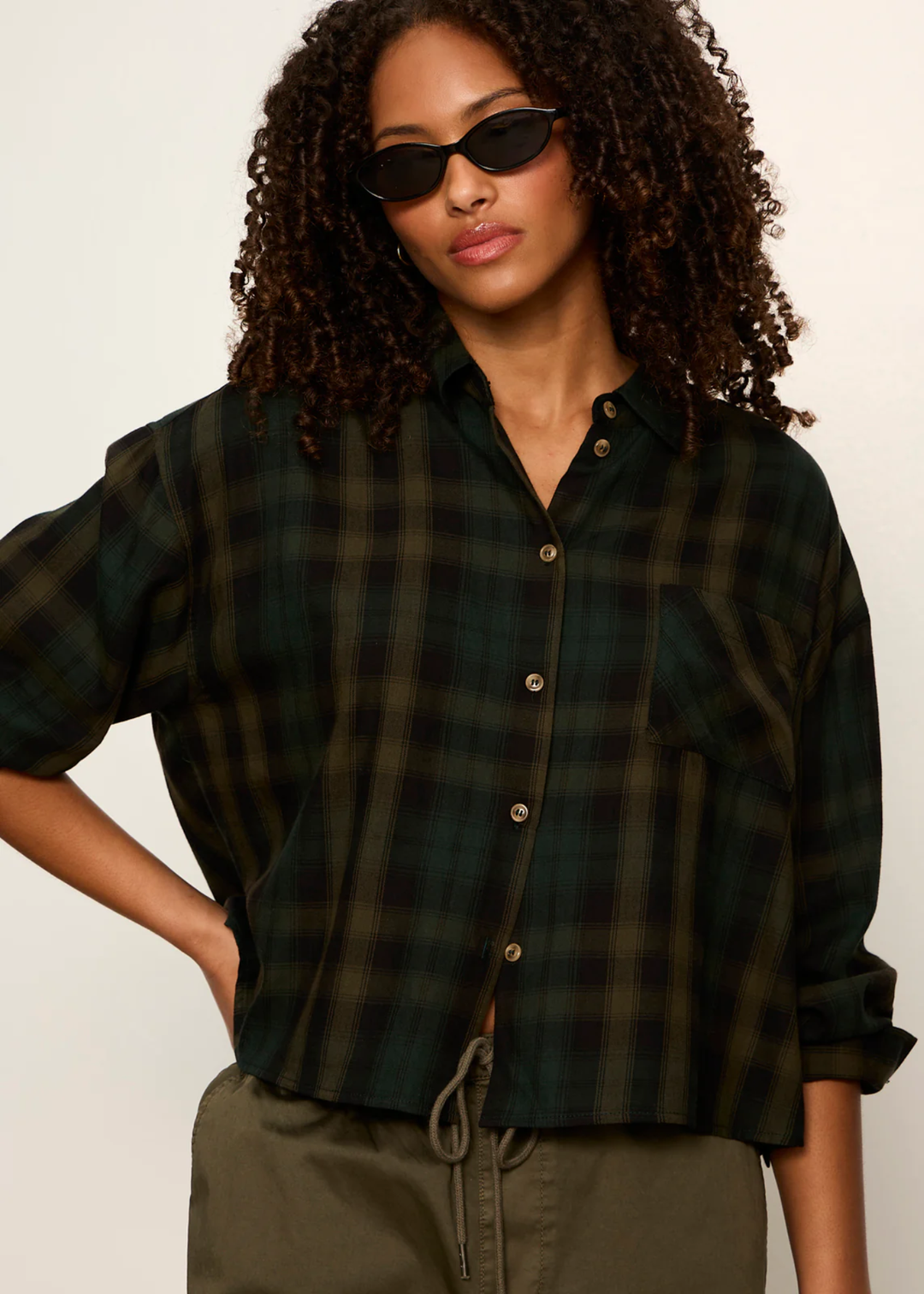 Woman wth curly hair and sunglasses wearing a forest plaid button down with hand on her hip.