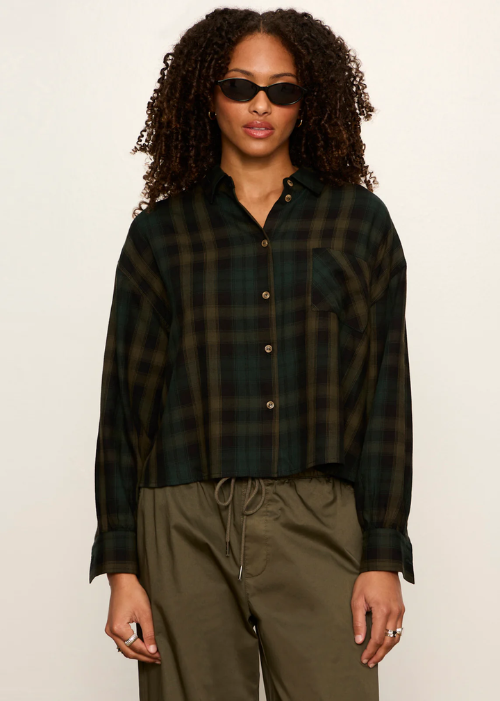 A model with curly hair wearing a dark green and black plaid button-up shirt and olive green drawstring pants, standing against a plain off-white background.