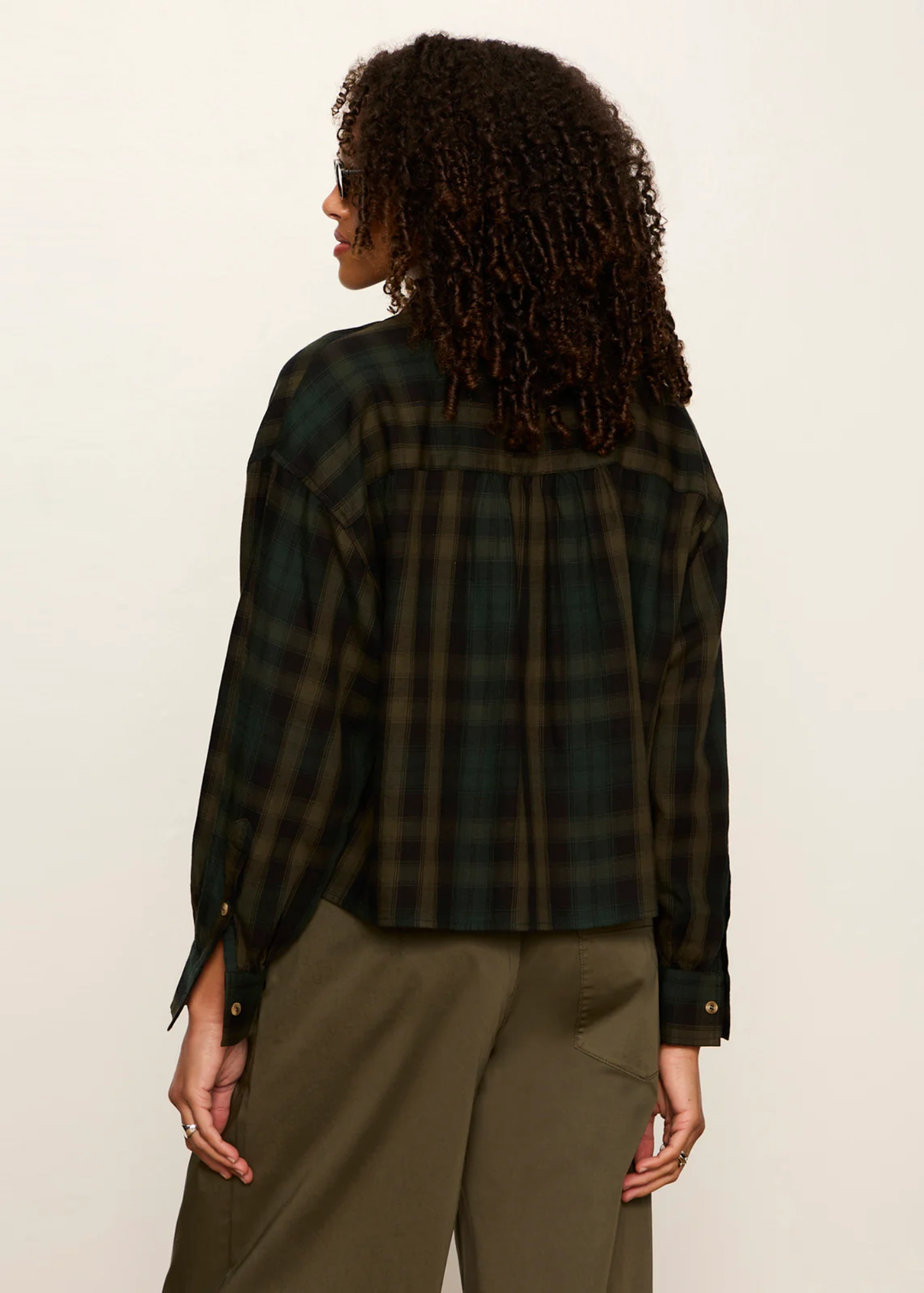 A woman shown from the back wearing a dark green and black plaid long-sleeve shirt with olive green pants, photographed against a plain off-white studio background.