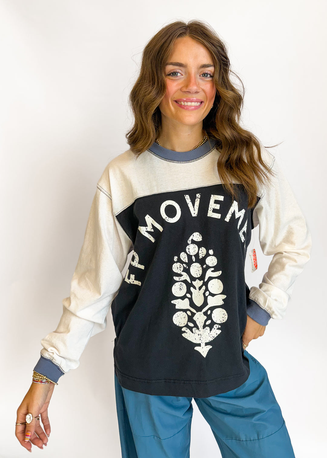 A woman modeling a long-sleeve crewneck sweatshirt with a color-blocked design. The top half is cream, the bottom half is black, and there is a white graphic print of a tree with fruit and the text 'FPMOVEMENT' across the chest.