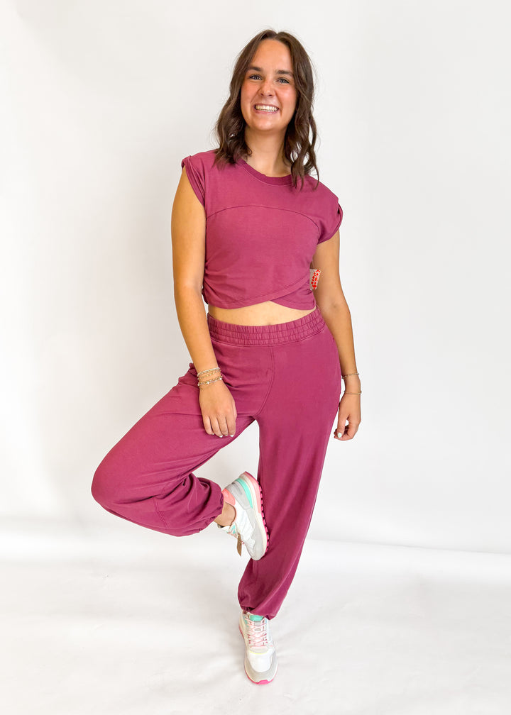 A woman modeling a matching two-piece lounge set consisting of a short-sleeve crop top and jogger pants in a dusty rose or mauve color, paired with white sneakers, against a plain white studio background.