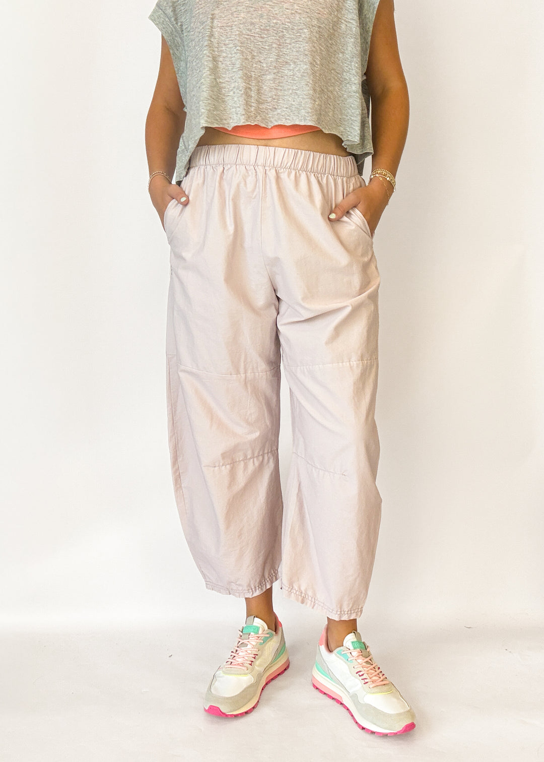 A person wearing loose, light blush-beige cropped pants with an elastic waistband and side pockets, paired with a gray sleeveless crop top and pastel sneakers, standing against a plain light background.
