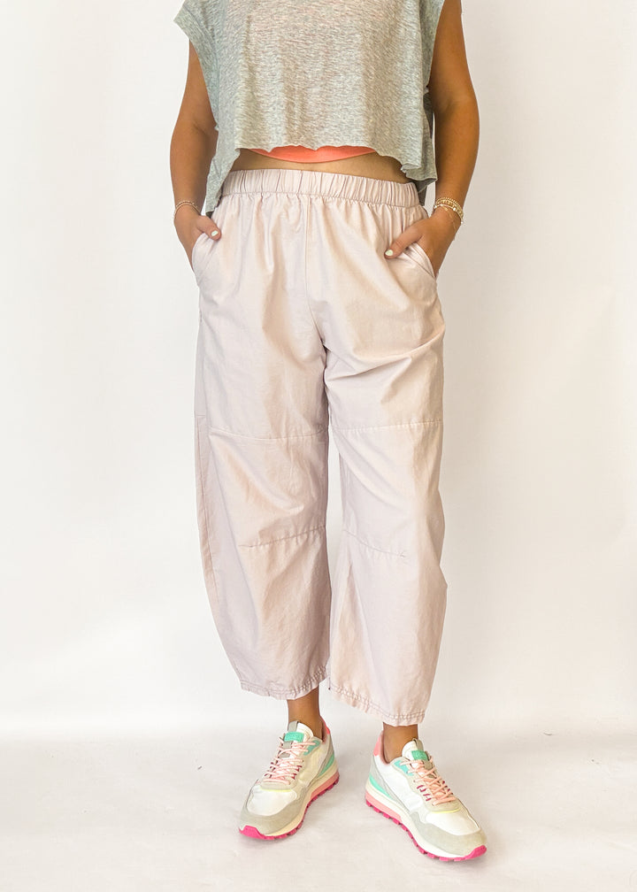 A person wearing loose, light blush-beige cropped pants with an elastic waistband and side pockets, paired with a gray sleeveless crop top and pastel sneakers, standing against a plain light background.