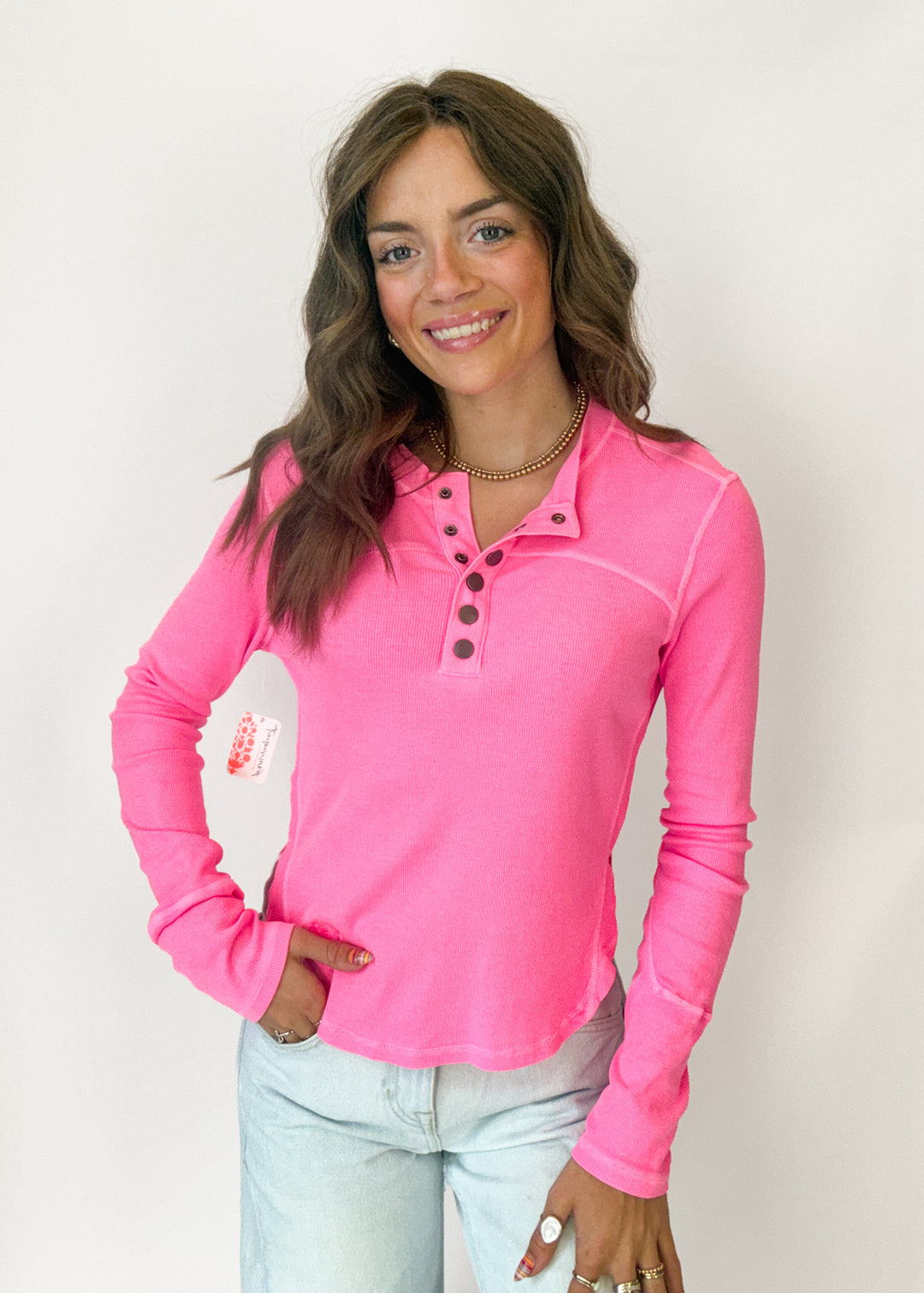 A woman modeling a bright pink long-sleeve henley shirt with a buttoned collar, paired with light blue jeans, against a plain white background.