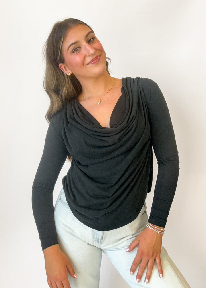 A woman modeling a black long-sleeve cowl neck top with draped fabric, paired with light-wash jeans, against a plain white background.