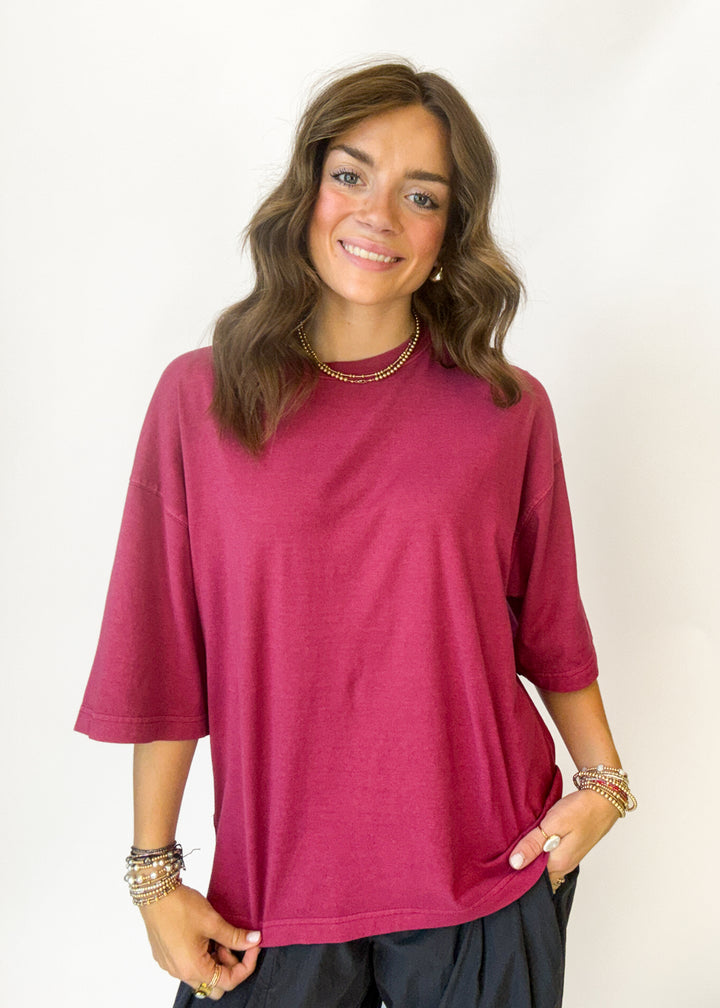 A woman wearing a relaxed-fit, three-quarter sleeve magenta top and dark pants, standing against a plain white background.