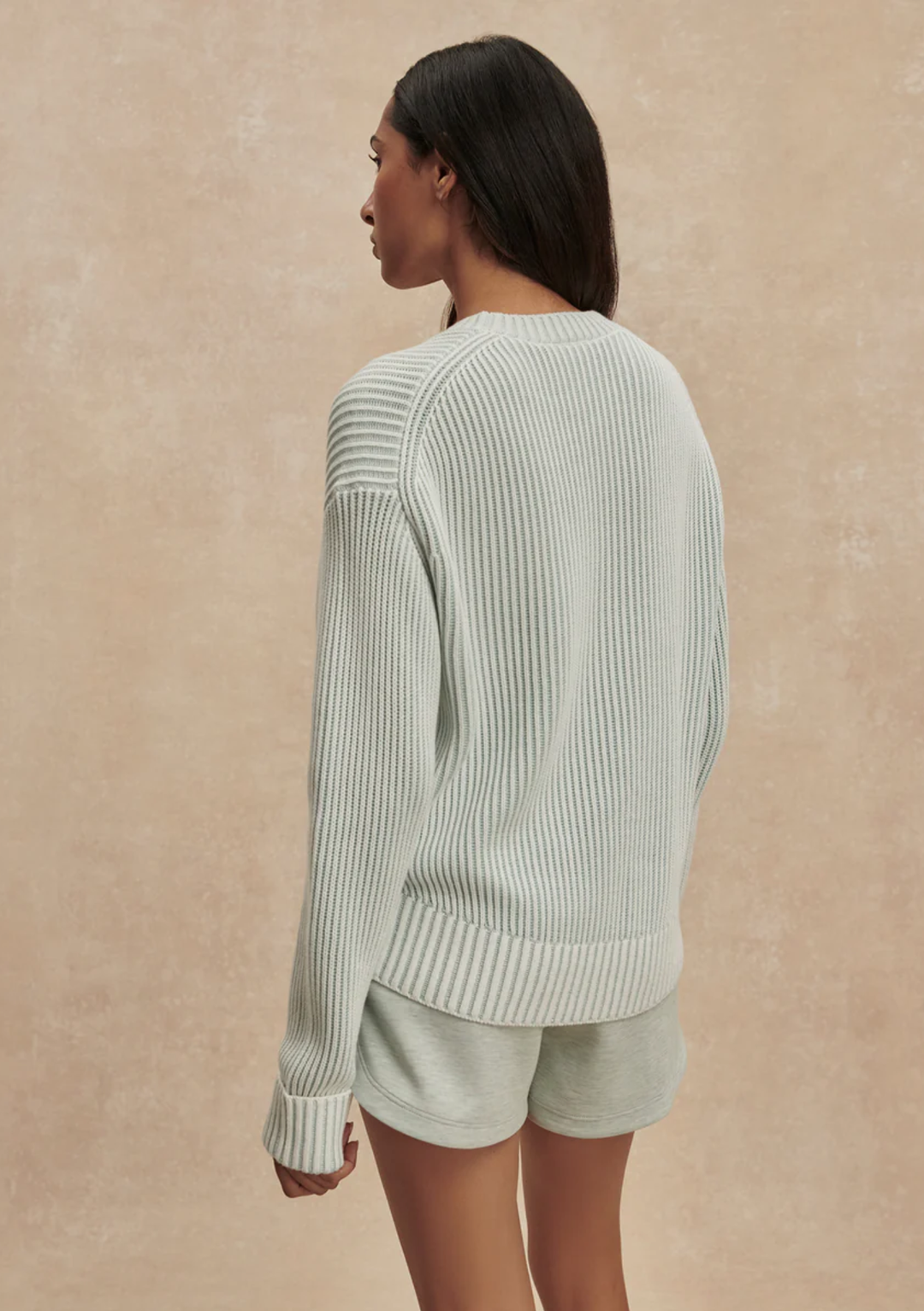 A woman shown from the back wearing a ribbed knit long-sleeve sweater and matching shorts in a pale sage green, set against a textured beige studio backdrop.