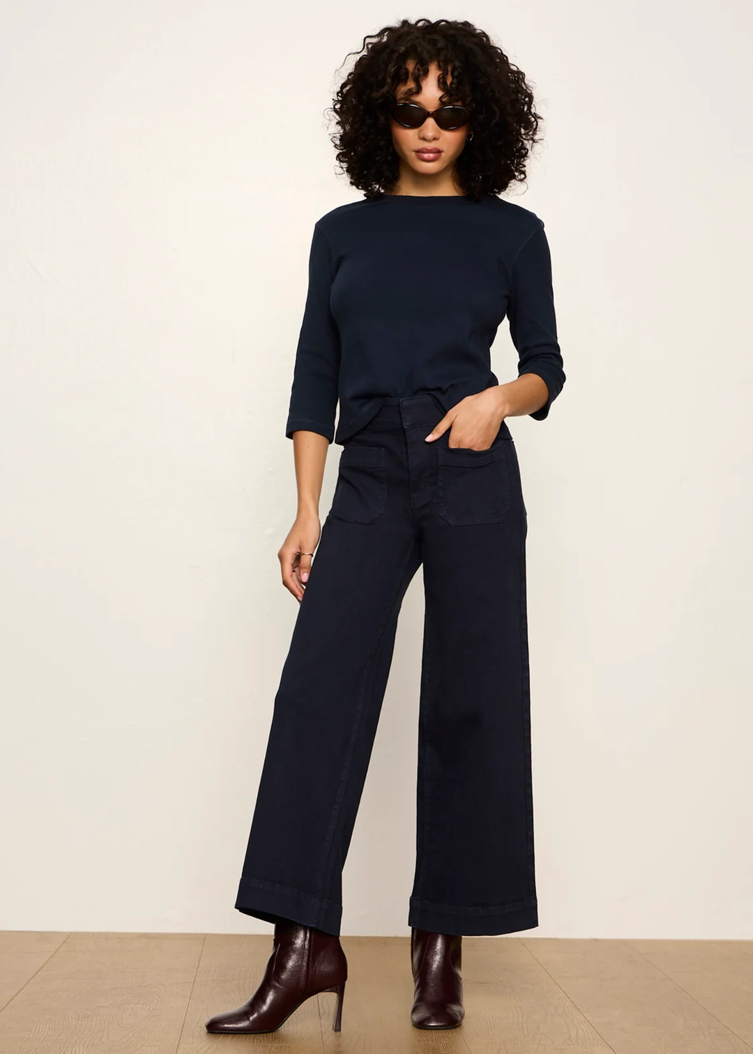 A woman modeling a dark navy blue three-quarter sleeve top and matching wide-leg trousers with a tie waist, styled with dark brown heeled ankle boots against a plain off-white studio background.
