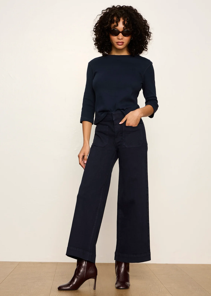 A woman modeling a dark navy blue three-quarter sleeve top and matching wide-leg trousers with a tie waist, styled with dark brown heeled ankle boots against a plain off-white studio background.